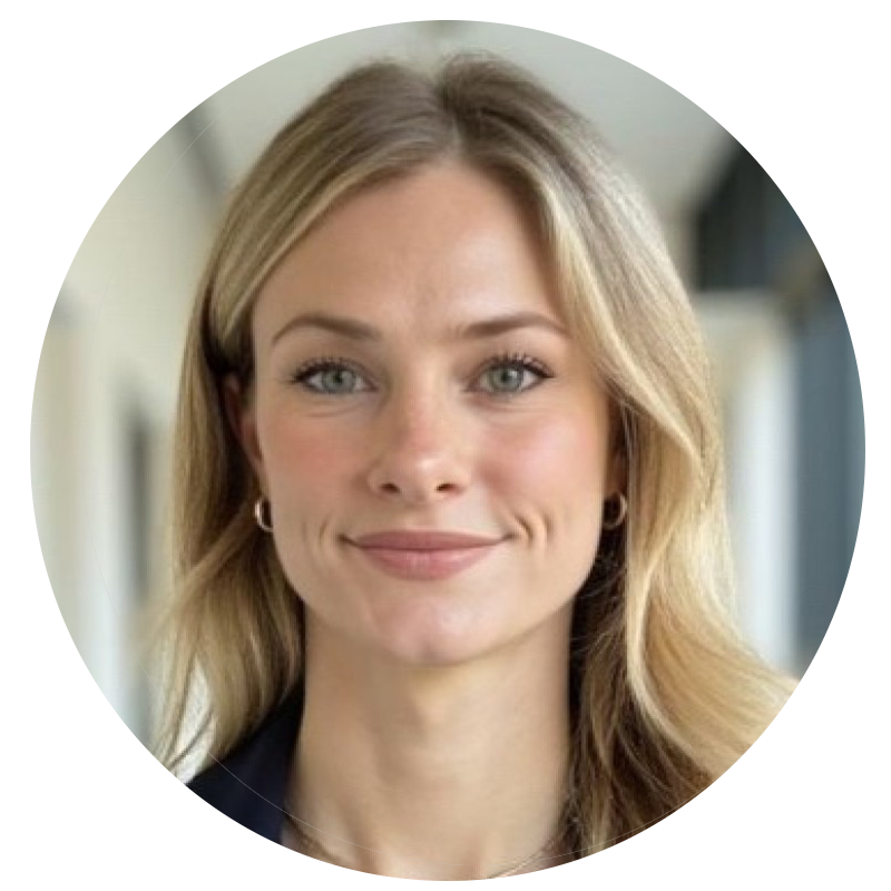 صورة شخصية احترافية لامرأة شقراء مبتسمة ترتدي سترة داكنة، وهي تقف في ردهة بالقرب من المكاتب.