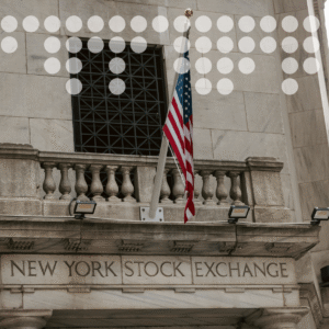 Η πρόσοψη του κτιρίου του Χρηματιστηρίου της Νέας Υόρκης, με μια αμερικανική σημαία να κυματίζει σε ένα πέτρινο μπαλκόνι, ένα μεγάλο παράθυρο με κάγκελα από πάνω και κλασικά αρχιτεκτονικά στοιχεία.