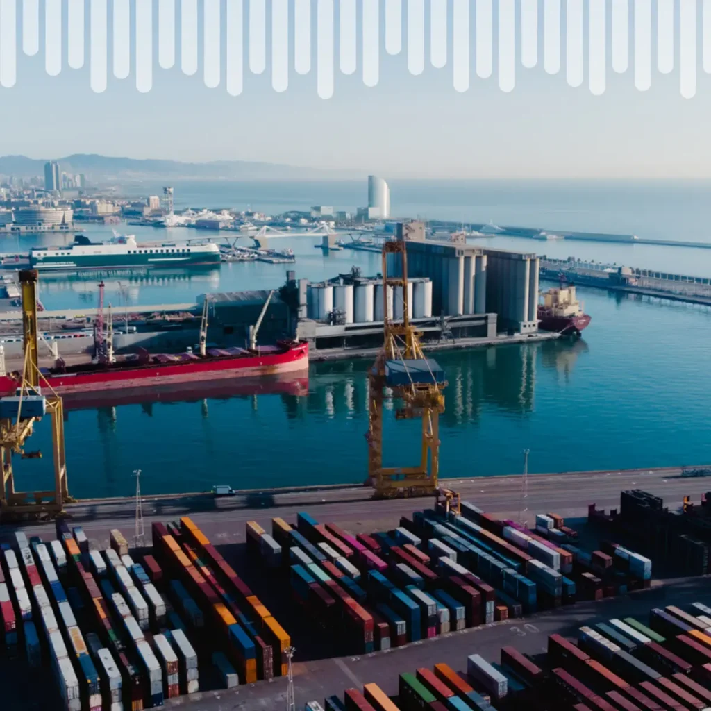 Wide view of a busy harbor with cargo ships, yellow cranes, and multicolored shipping containers along the dock in calm water.