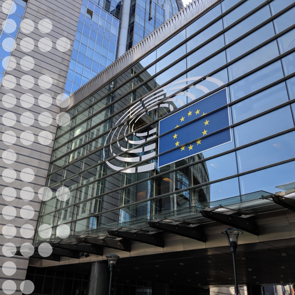 Edificio de oficinas acristalado con la bandera de la Unión Europea en la fachada, en la que se refleja el cielo azul, con un motivo arquitectónico circular en el lado izquierdo.