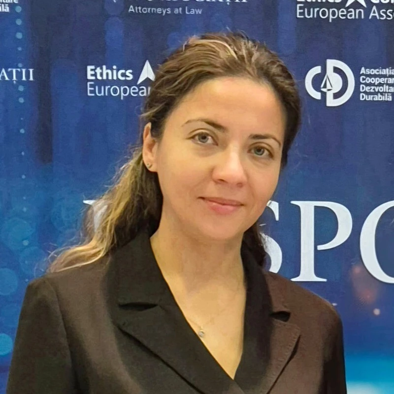 Eine berufstätige Frau in einem schwarzen Blazer posiert vor einem blauen Konferenzhintergrund, hinter ihr sind Logos und Schriftzüge zu sehen