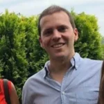 Uomo sorridente con una camicia azzurra all’aperto, con delle siepi verdi sullo sfondo.
