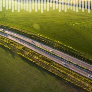 Vue aérienne d'une autoroute à plusieurs voies traversant des terres agricoles verdoyantes, avec des rangées d'arbres le long du terre-plein central et, au loin, des champs aux contours sinueux.